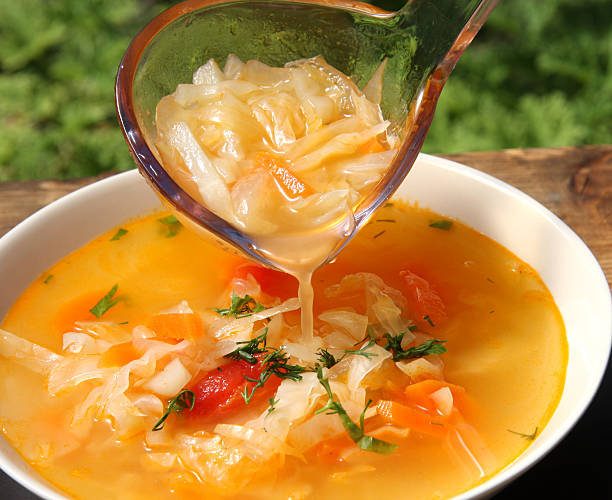 A ladle serving cabbage soup from a bowl on a wooden table, surrounded by greenery.