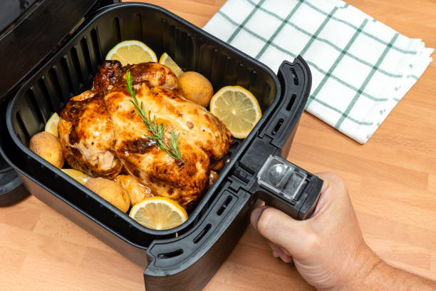 Hand holding an air fryer basket containing a cooked chicken surrounded by potatoes and lemon slices.