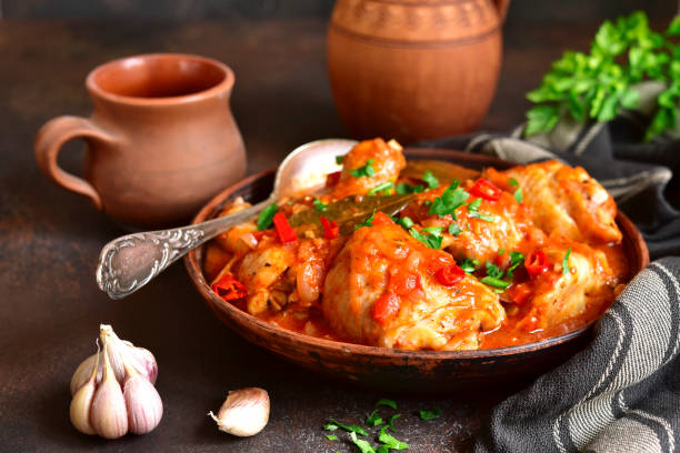 A bowl of chicken stew garnished with herbs and peppers, surrounded by garlic bulbs and a clay cup.