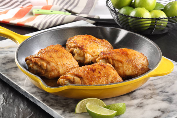 Four golden-brown chicken thighs in a yellow skillet on a marble surface, with lime wedges and a bowl of limes in the background.