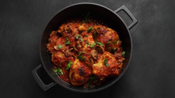 A black pot filled with chicken thighs in a rich tomato sauce, garnished with fresh parsley and mushrooms.