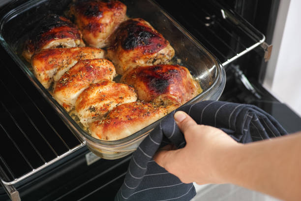 A hand wearing a striped oven mitt is pulling a glass baking dish filled with golden brown roasted chicken from an oven.