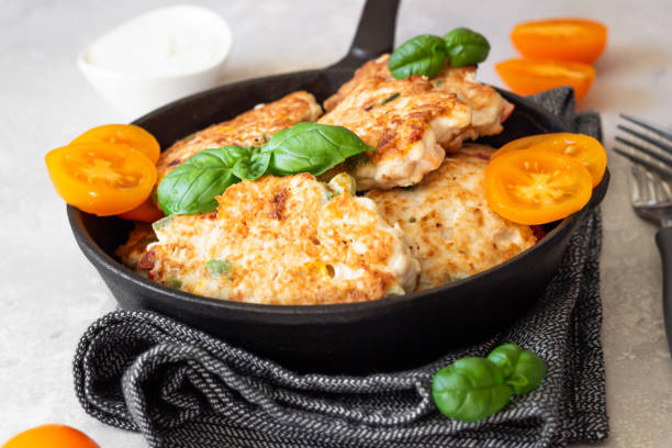 A cast iron skillet filled with golden brown vegetable fritters adorned with fresh basil leaves and sliced yellow tomatoes.