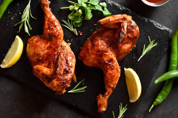 Two pieces of well-cooked spicy roast chicken on a black slate plate with fresh herbs and lemon wedges