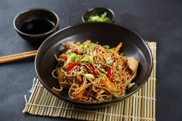 A bowl of stir-fried noodles with vegetables and chicken, garnished with green onions and sesame seeds, served on a bamboo mat.