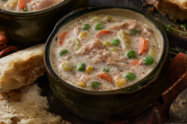 A close-up of a bowl of creamy chicken soup with vegetables, surrounded by pieces of bread.