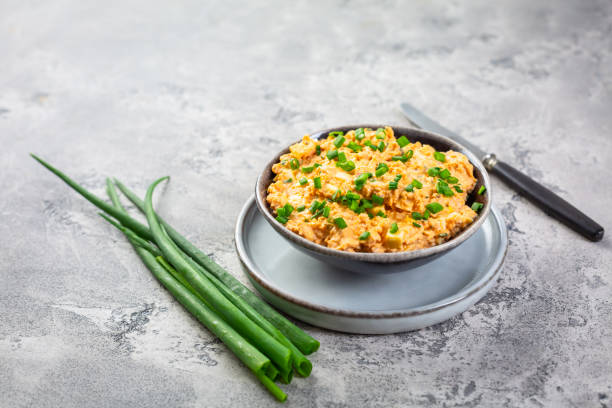 A bowl of creamy potato salad topped with chopped green onions, sitting on a grey stone surface next to fresh green onion stalks and a knife.