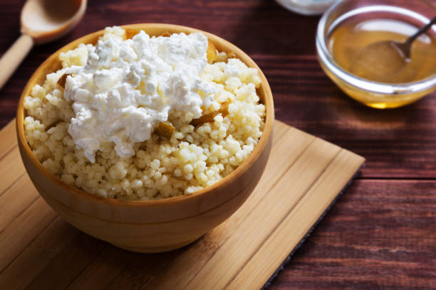 A wooden bowl filled with couscous topped with a generous serving of cottage cheese, sitting on a wooden cutting board next to a small bowl of honey.