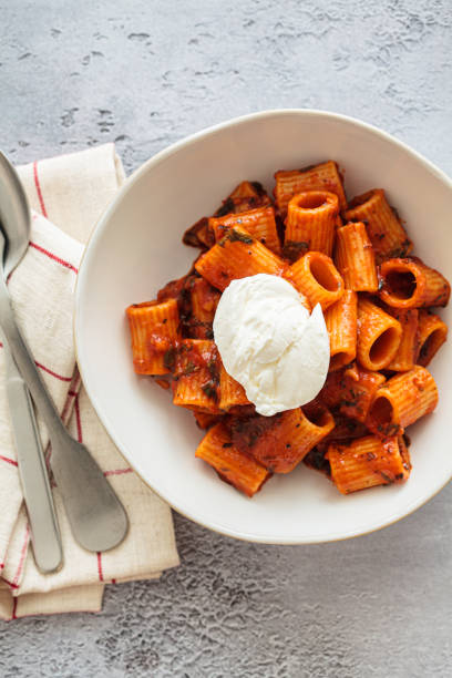 A bowl of rigatoni pasta topped with a dollop of cream, served on a light background with utensils beside it.