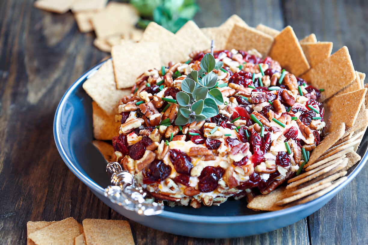 A colorful cheese ball topped with nuts and cranberries, surrounded by crispy crackers.