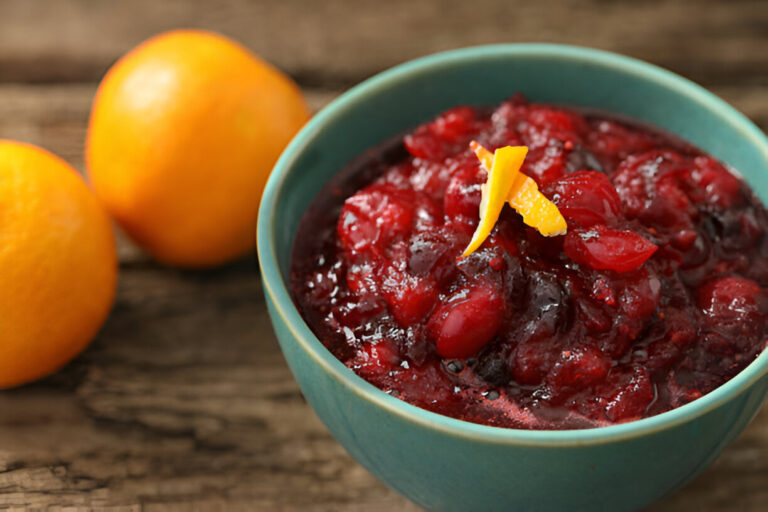 A bowl of cranberry sauce garnished with orange peel, accompanied by two oranges on a wooden surface.