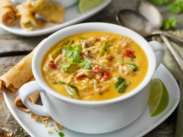 A bowl of creamy soup with vegetables and herbs, accompanied by spring rolls and a slice of lime.