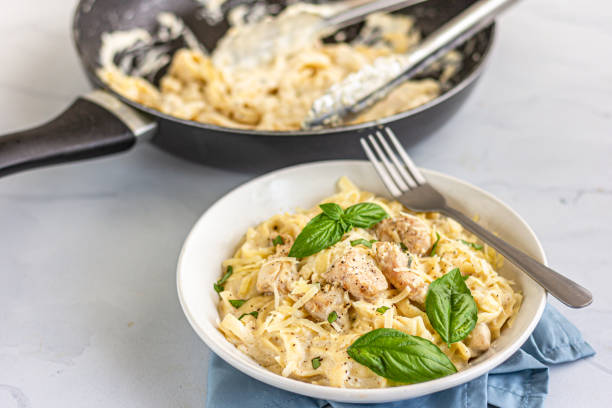 A bowl of creamy fettuccine pasta with chicken and fresh basil on a light-colored surface, next to a frying pan with pasta remnants.