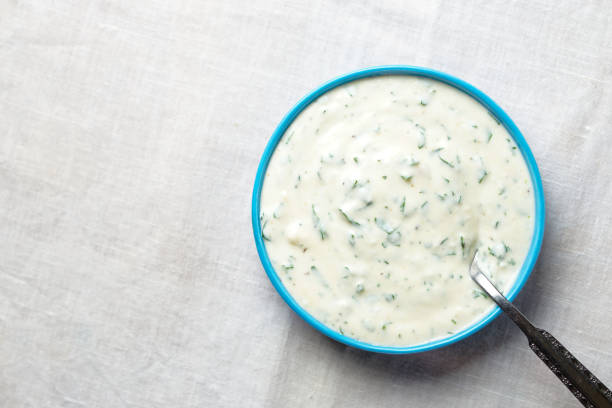 A bowl of creamy sauce with herbs and a spoon on a textured surface