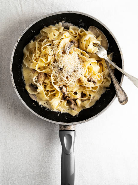 A pan filled with creamy fettuccine pasta topped with cheese and mushrooms, resting on a white surface.