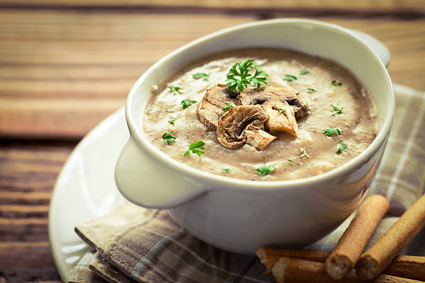 A bowl of creamy mushroom soup garnished with chopped parsley and mushrooms, placed on a wooden table.