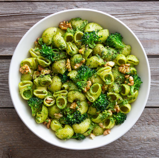 A bowl of green pasta with broccoli and walnuts on a wooden table.