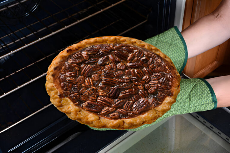 A person wearing green oven mitts is holding a freshly baked pecan pie just taken out of the oven.