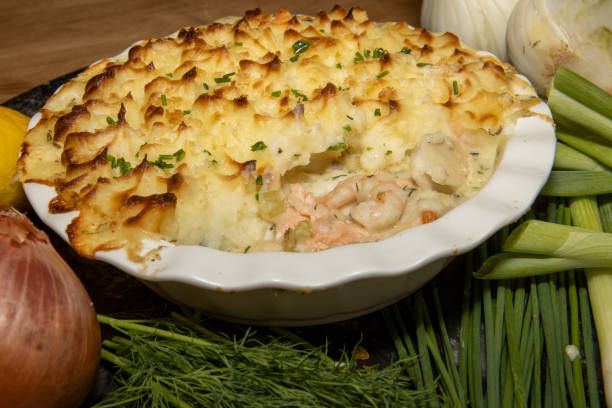 A baked fish pie topped with golden mashed potatoes, surrounded by fresh ingredients including an onion, lemon, and green herbs.