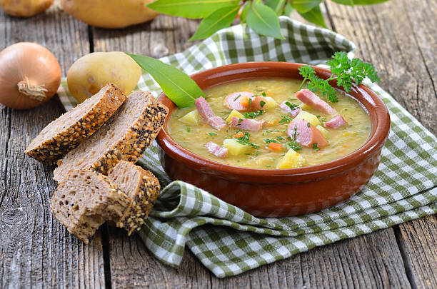 A bowl of hearty soup with pieces of meat and vegetables next to slices of dark bread on a checkered cloth.