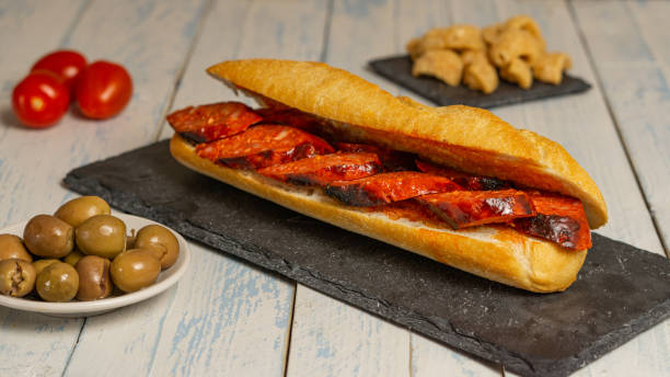 A delicious sandwich filled with sliced chorizo on a rustic wooden table, accompanied by olives, cherry tomatoes, and crispy snacks.