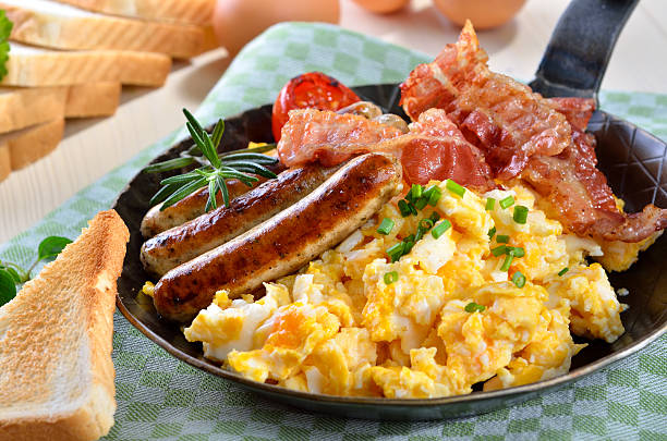 A delicious breakfast platter featuring scrambled eggs, sausages, crispy bacon, a grilled tomato, and slices of toast on a checkered cloth.