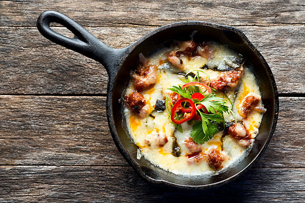 A cast iron skillet filled with creamy baked dish topped with herbs and red chili slices on a rustic wooden table.