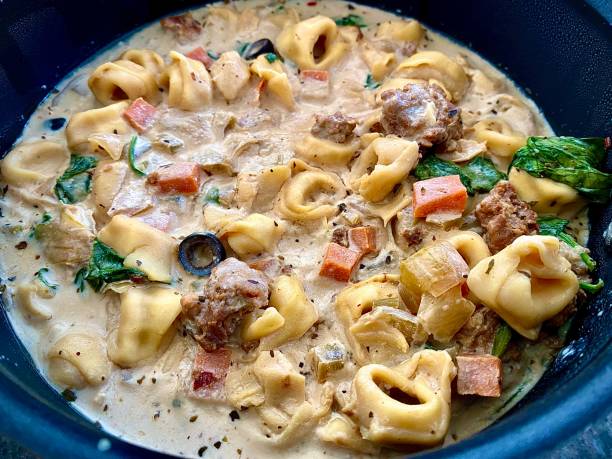 A creamy tortellini soup with meatballs, spinach, and diced vegetables in a black bowl.