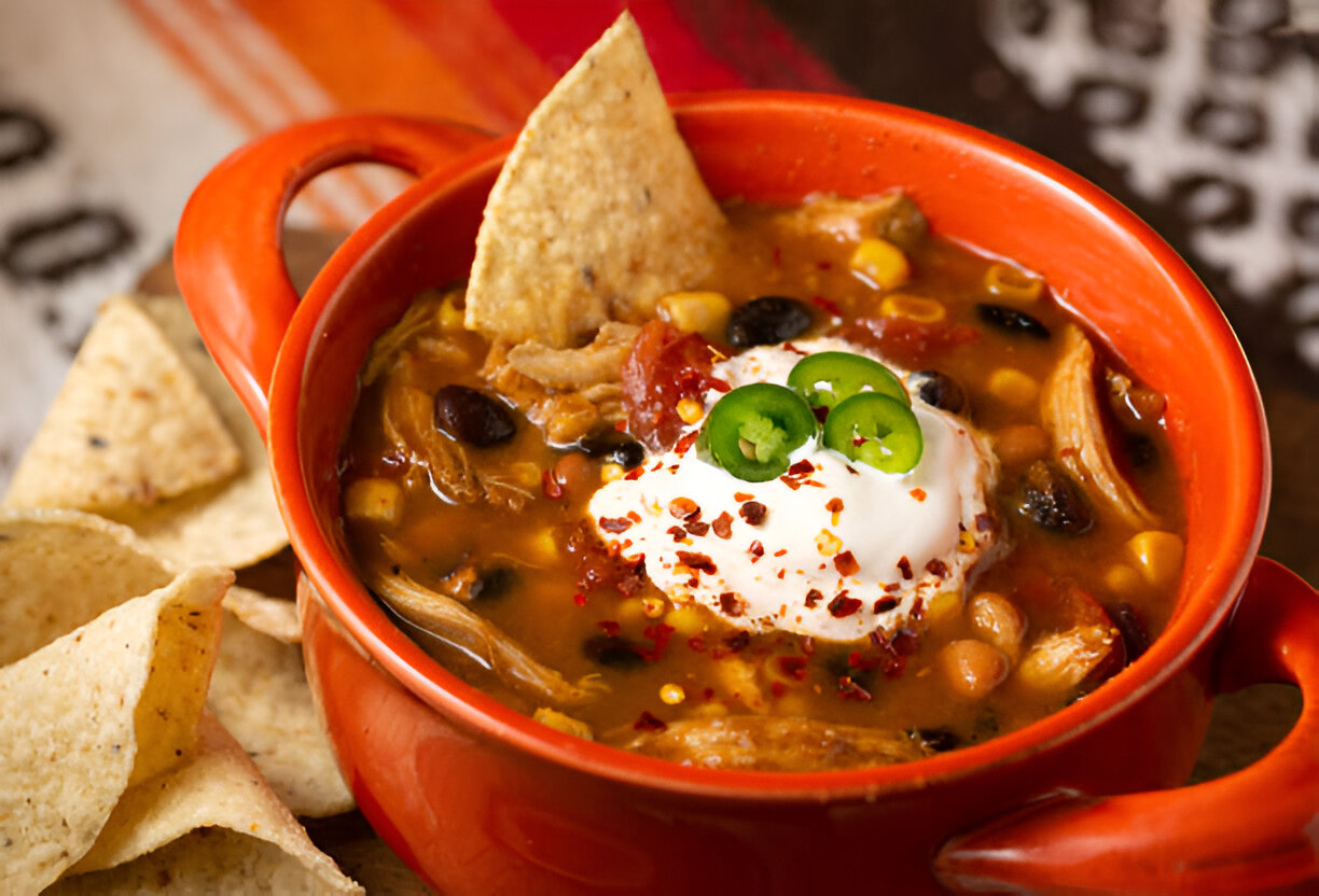 A vibrant bowl of chicken tortilla soup topped with sour cream, jalapeño slices, and crushed red pepper, alongside tortilla chips.