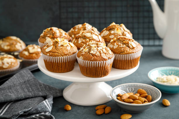 A display of muffins topped with sliced almonds on a white cake stand, accompanied by a small bowl of almonds and a blue decorative plate.