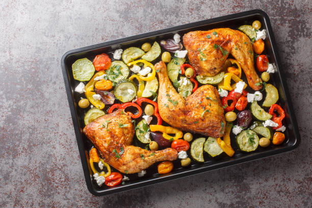 A tray of roasted chicken legs served with assorted vegetables including peppers, olives, and zucchini.