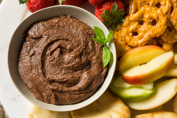 A bowl of chocolate dip surrounded by fresh strawberries, apple slices, and pretzels.