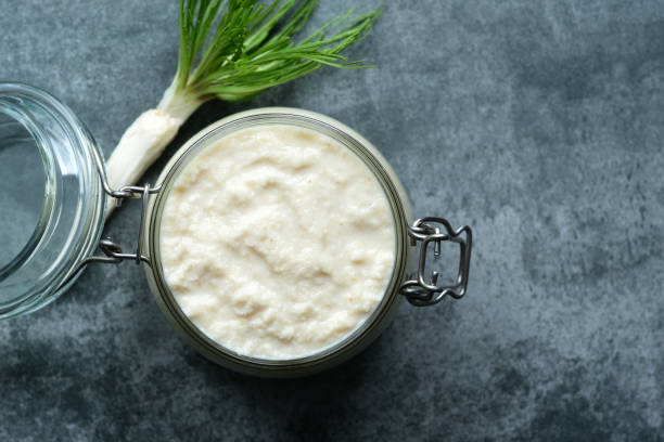 A jar of creamy white condiment with a sprig of green herbs beside it on a textured gray surface.