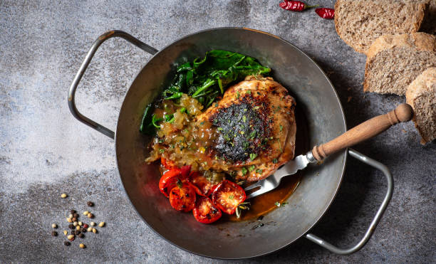 A delicious plate of grilled chicken with sautéed spinach, roasted tomatoes, and a side of bread, served in a metal skillet on a rustic surface.