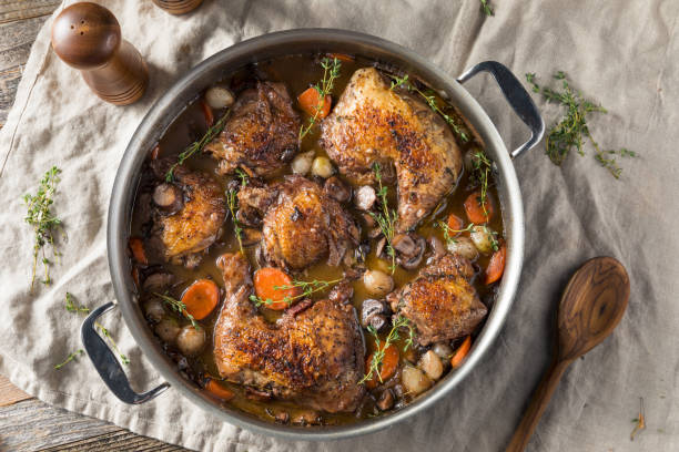 A pot of braised chicken thighs surrounded by vegetables and herbs sitting on a linen cloth.