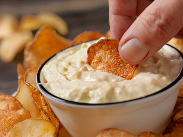 A hand dipping a potato chip into a bowl of creamy dip, surrounded by chips.