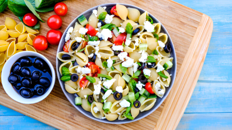 A colorful pasta salad with shell pasta, cherry tomatoes, green peppers, black olives, and cheese, served on a wooden cutting board.