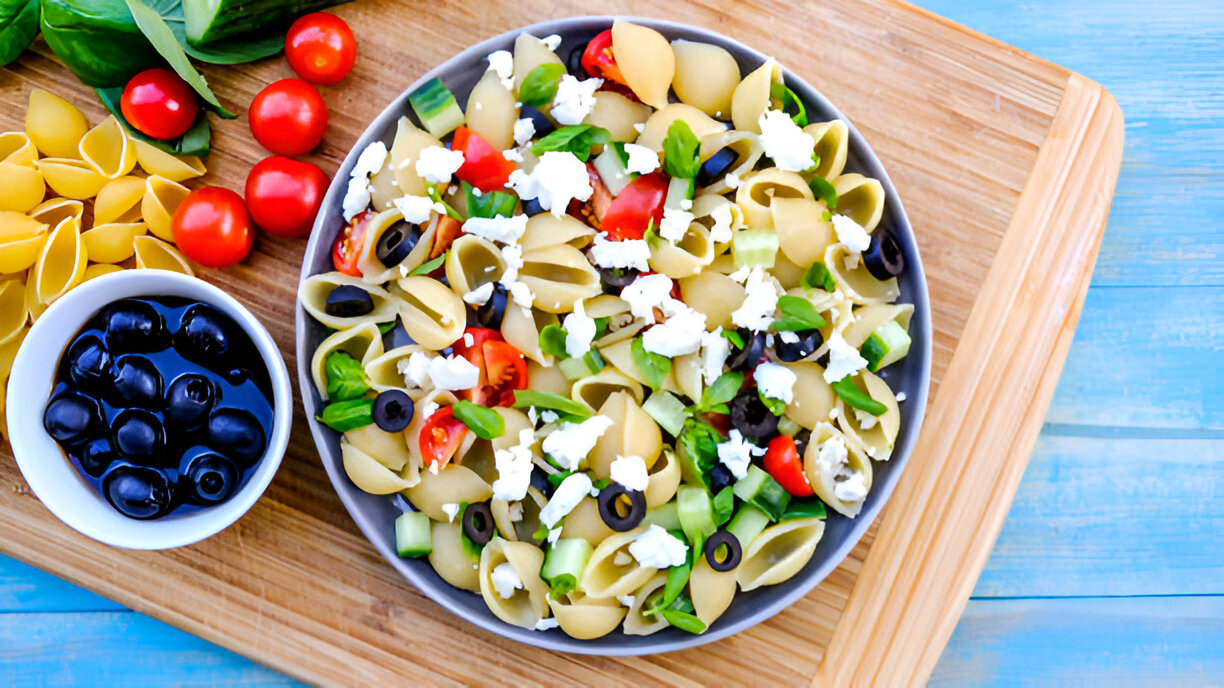 A colorful pasta salad with shell pasta, cherry tomatoes, green peppers, black olives, and cheese, served on a wooden cutting board.