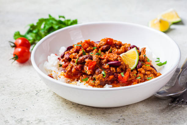 A bowl of chili served over white rice with a lime wedge and garnished with herbs, accompanied by cherry tomatoes.