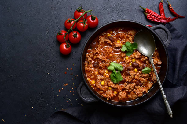 A delicious bowl of chili topped with fresh cilantro, served with tomatoes and red chili peppers on a dark background.