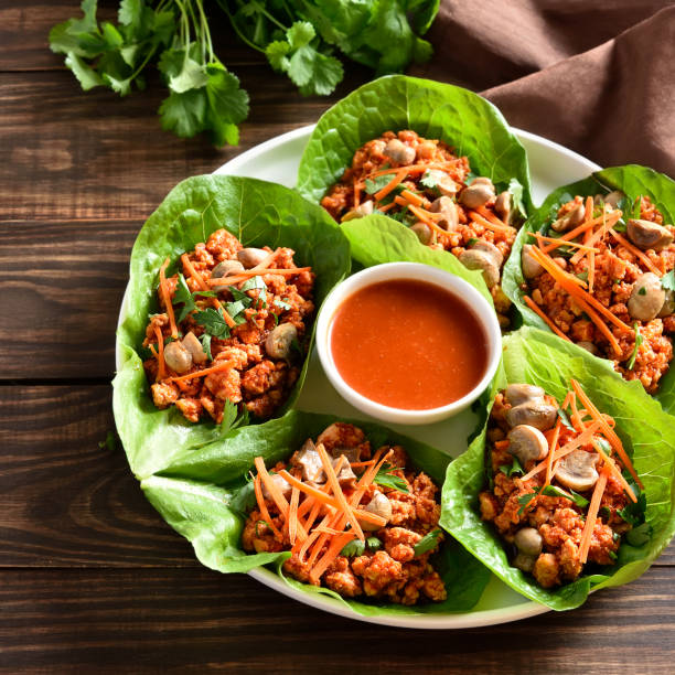 A platter of lettuce wraps filled with a spicy chicken mixture, garnished with carrots and cilantro, served with a small bowl of sauce.