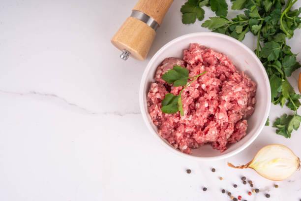 A bowl of freshly minced meat garnished with cilantro, surrounded by green herbs, a pepper grinder, and spices.