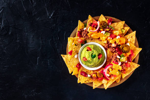 A wooden platter filled with golden nacho chips topped with various toppings and a bowl of guacamole in the center.