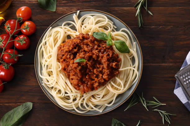 A plate of spaghetti with meat sauce, garnished with basil leaves, surrounded by fresh tomatoes and herbs.