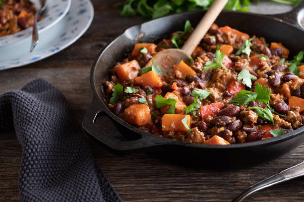 A skillet filled with chili featuring beans, pumpkin, and ground meat, garnished with fresh herbs.