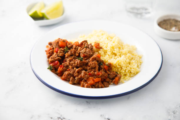 A plate of cooked ground meat with vegetables served alongside fluffy couscous, garnished with lime wedges.