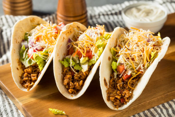 Three delicious tacos filled with ground meat, lettuce, tomatoes, cheese, and toppings on a wooden cutting board.