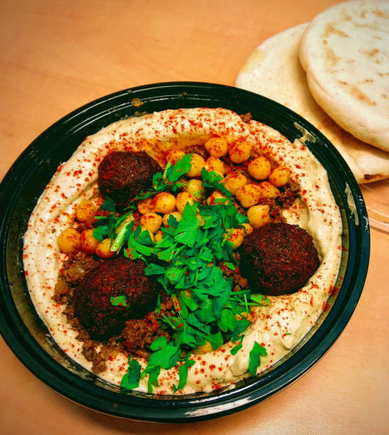 A bowl of hummus topped with chickpeas, parsley, and falafel balls, with pita bread on the side.