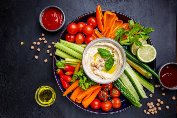 A vibrant platter of fresh vegetables including carrots, celery, cherry tomatoes, and bell peppers, accompanied by a bowl of creamy hummus garnished with olive oil and basil leaves.