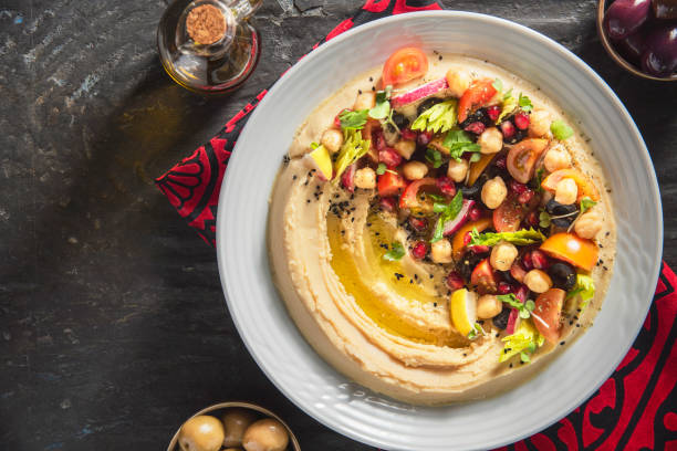 A bowl of hummus topped with colorful vegetables and herbs, drizzled with olive oil, placed on a dark stone surface.